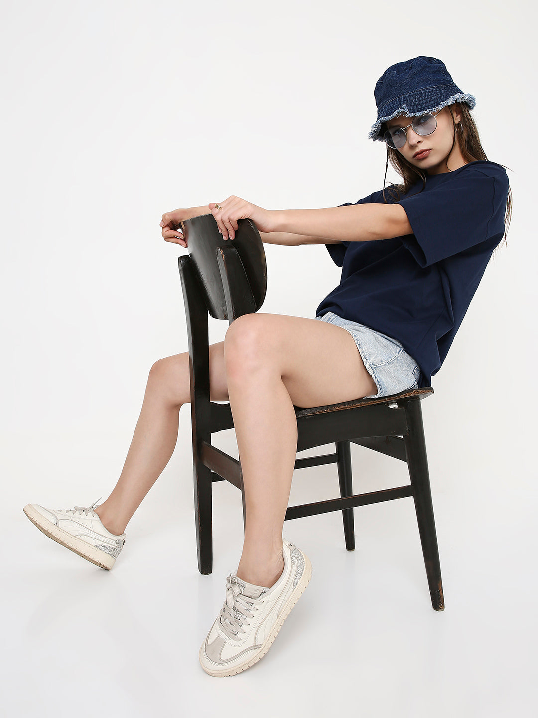 Woman wearing relaxed fit navy-blue solid t-shirt, denim shorts, and white sneakers sitting sideways on black wooden chair.
