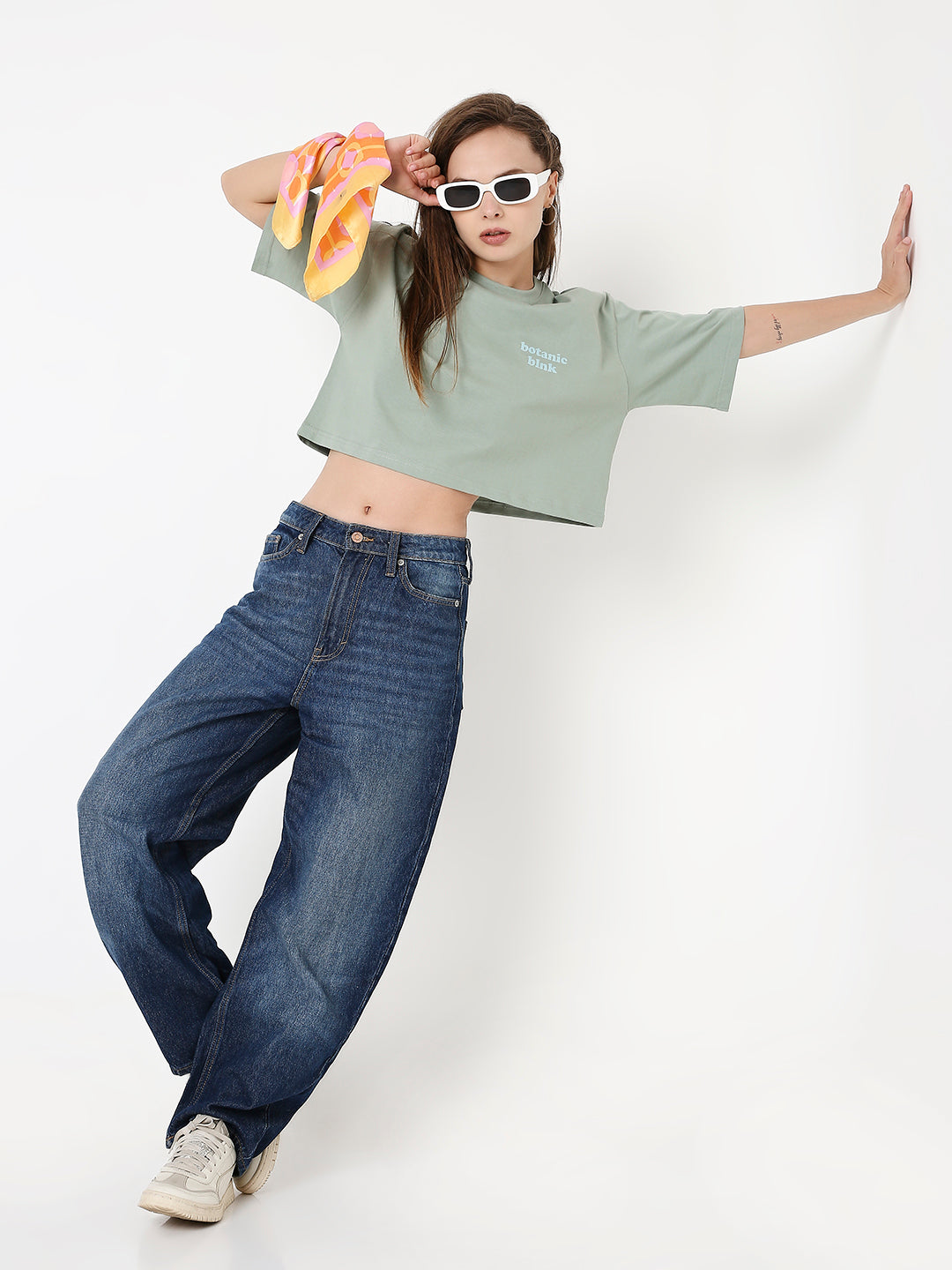 Oversize sage green crop top with chest print, paired with loose dark blue jeans and white sneakers.