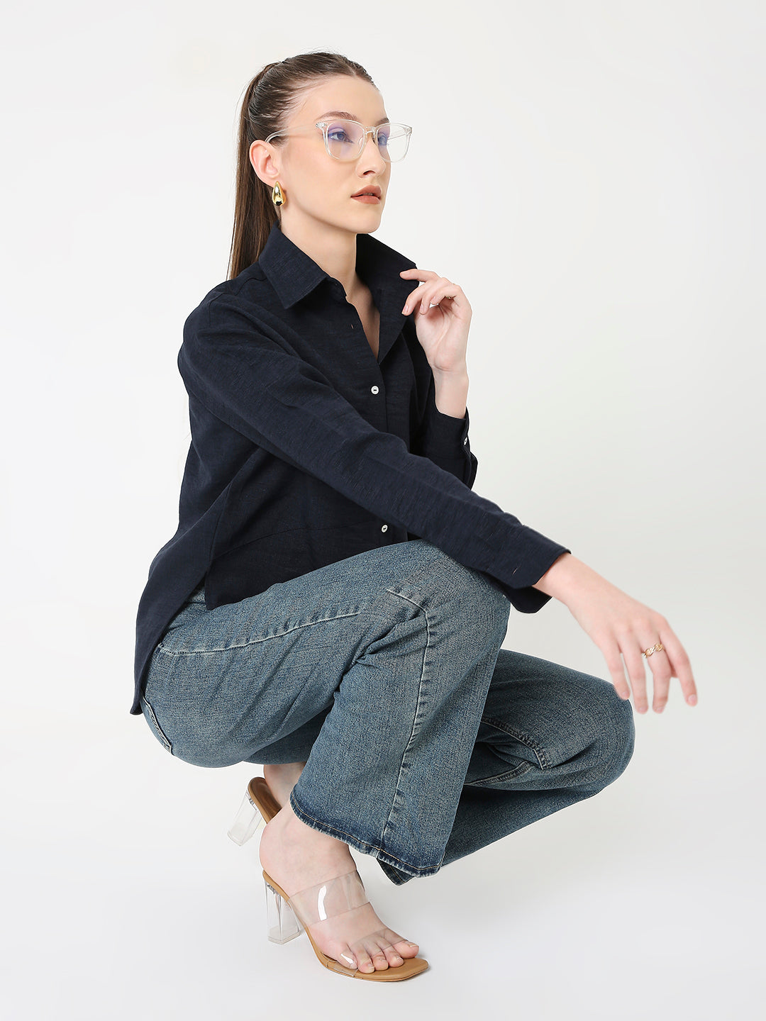 A woman is posing while wearing a dark navy button-down shirt with a classic collar, long sleeves, and front button-down closure. She is also wearing light blue jeans and beige heels.