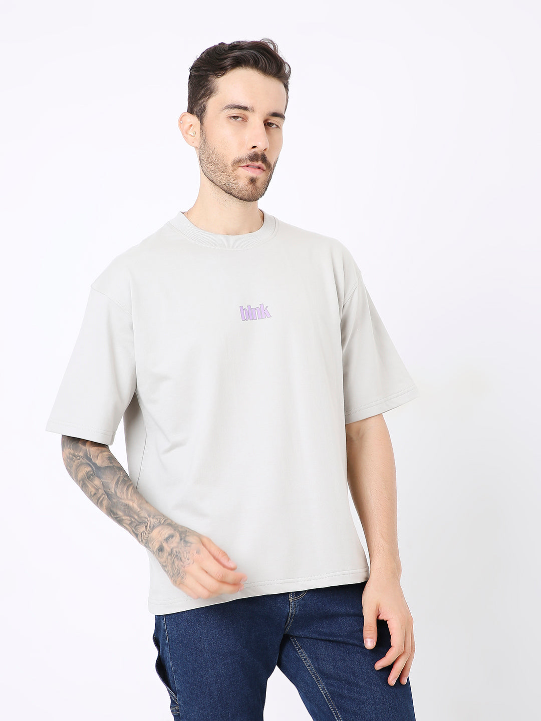 Light beige oversized t-shirt with small purple skipping stone graphic on chest, worn with dark jeans.