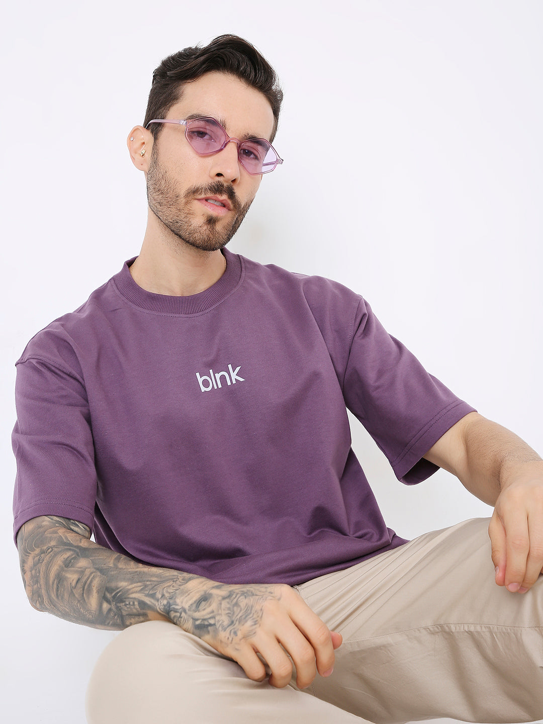 Oversize purple T-shirt with small white blink logo on chest, worn by tattooed man with glasses.