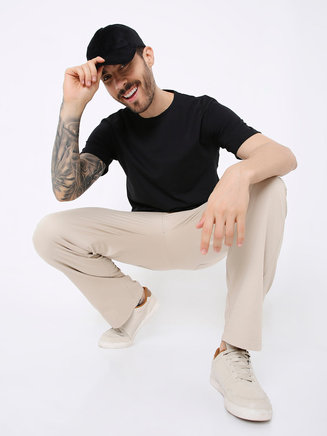 Man wearing relaxed fit black solid t-shirt, beige pants, white sneakers, and black cap, squatting.