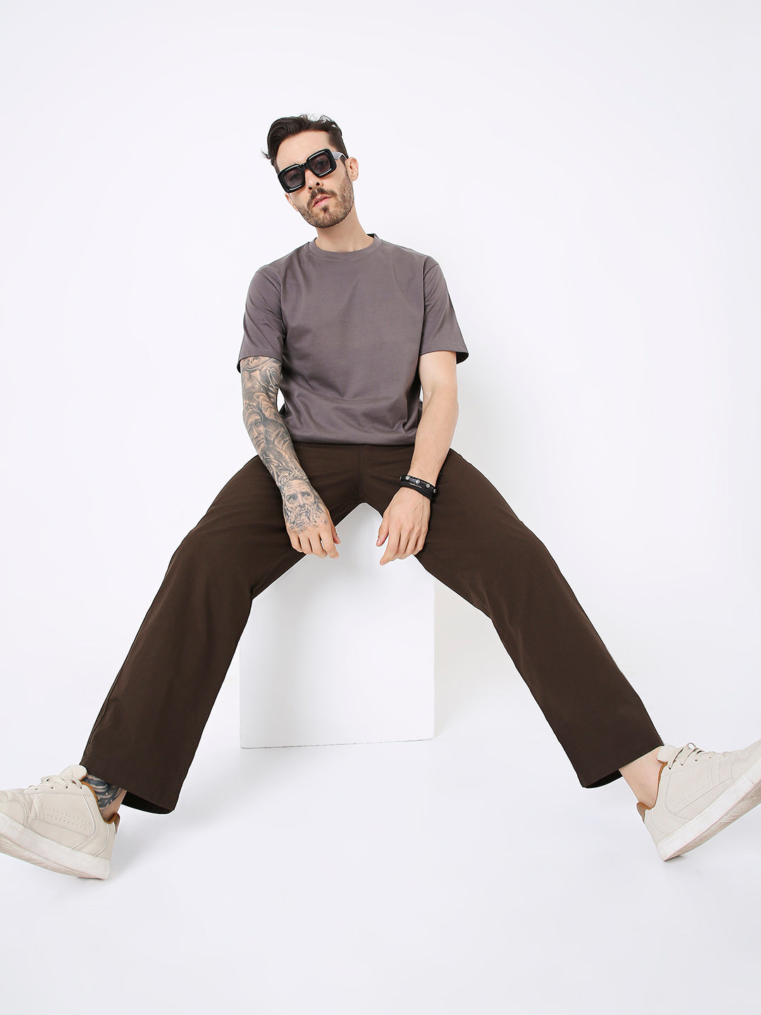 Man wearing relaxed fit steel grey solid t-shirt with dark brown wide-leg pants and beige sneakers sitting on white block.