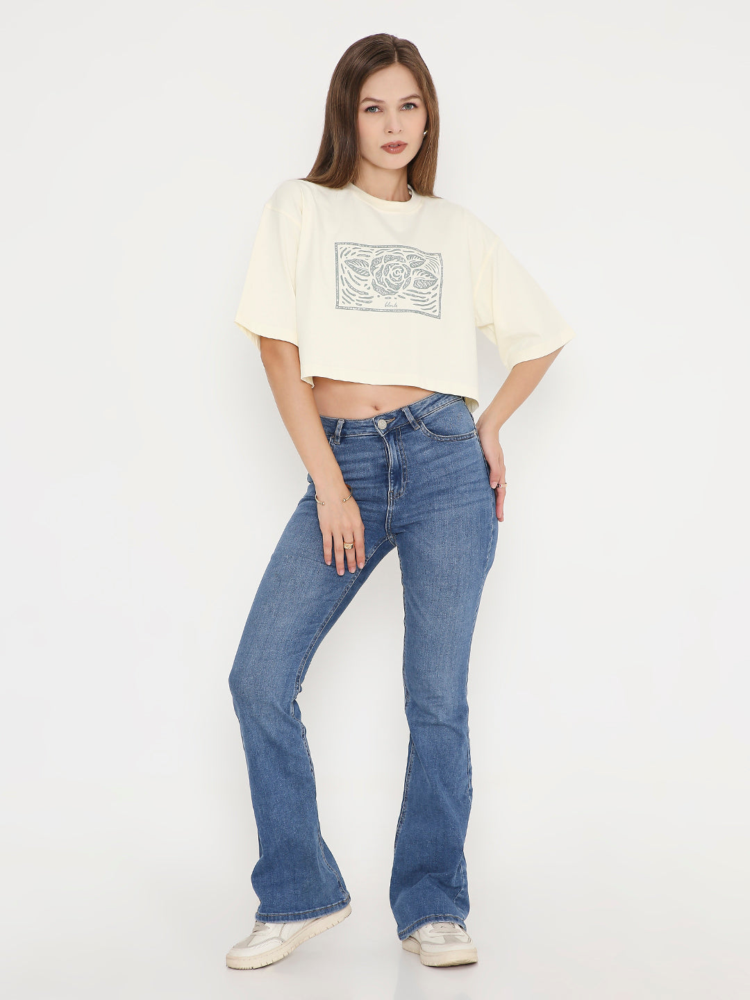 Off-white oversized crop top with abstract black print, paired with blue bootcut jeans and white sneakers.