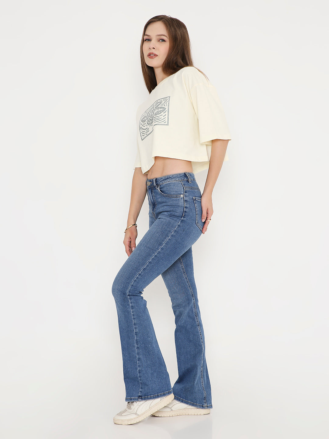 Off-white oversized crop top with black abstract print, paired with blue bootcut jeans and white sneakers.