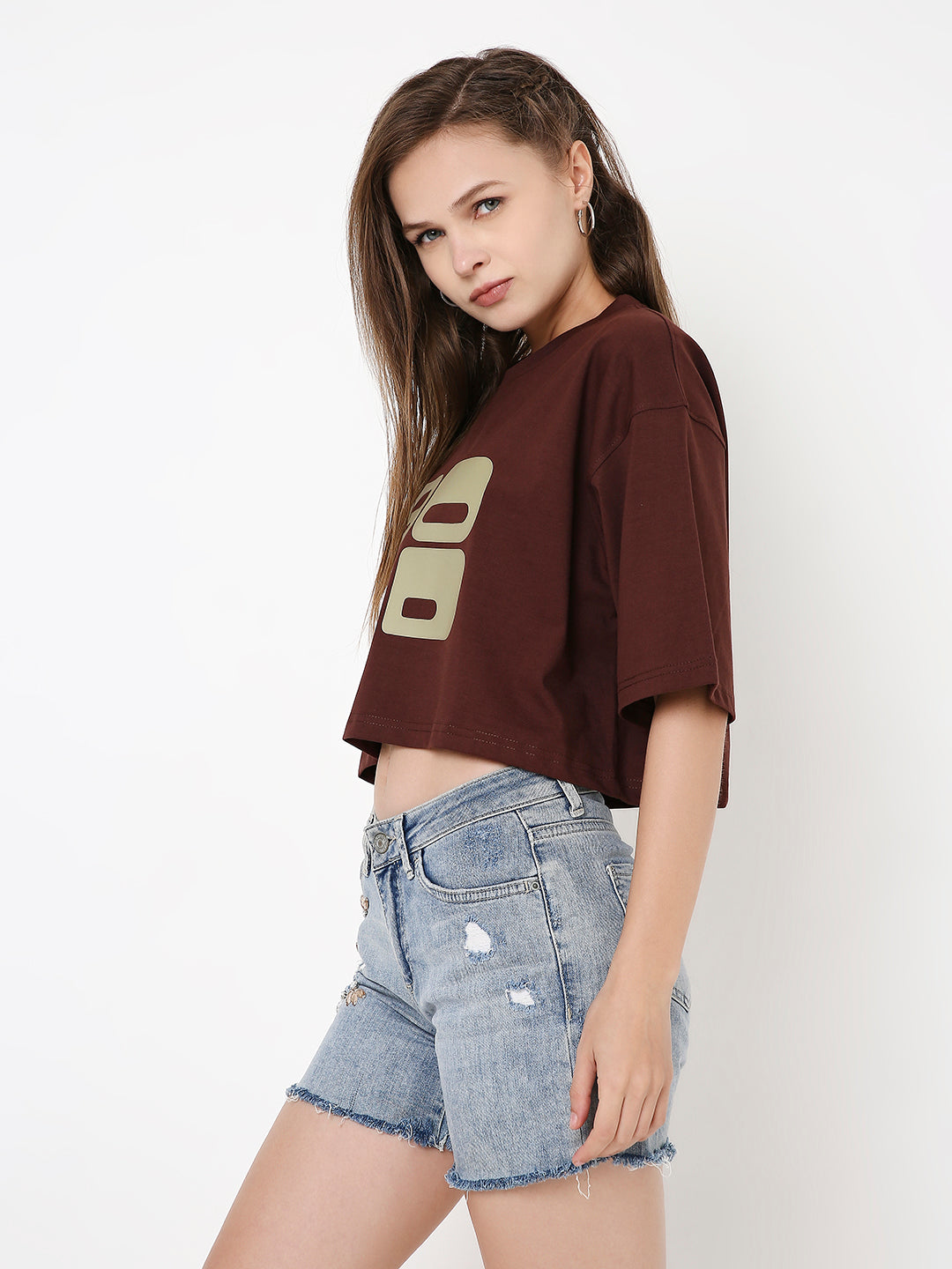 Oversize brown crop top with beige graphic print and dropped shoulders paired with distressed denim shorts.