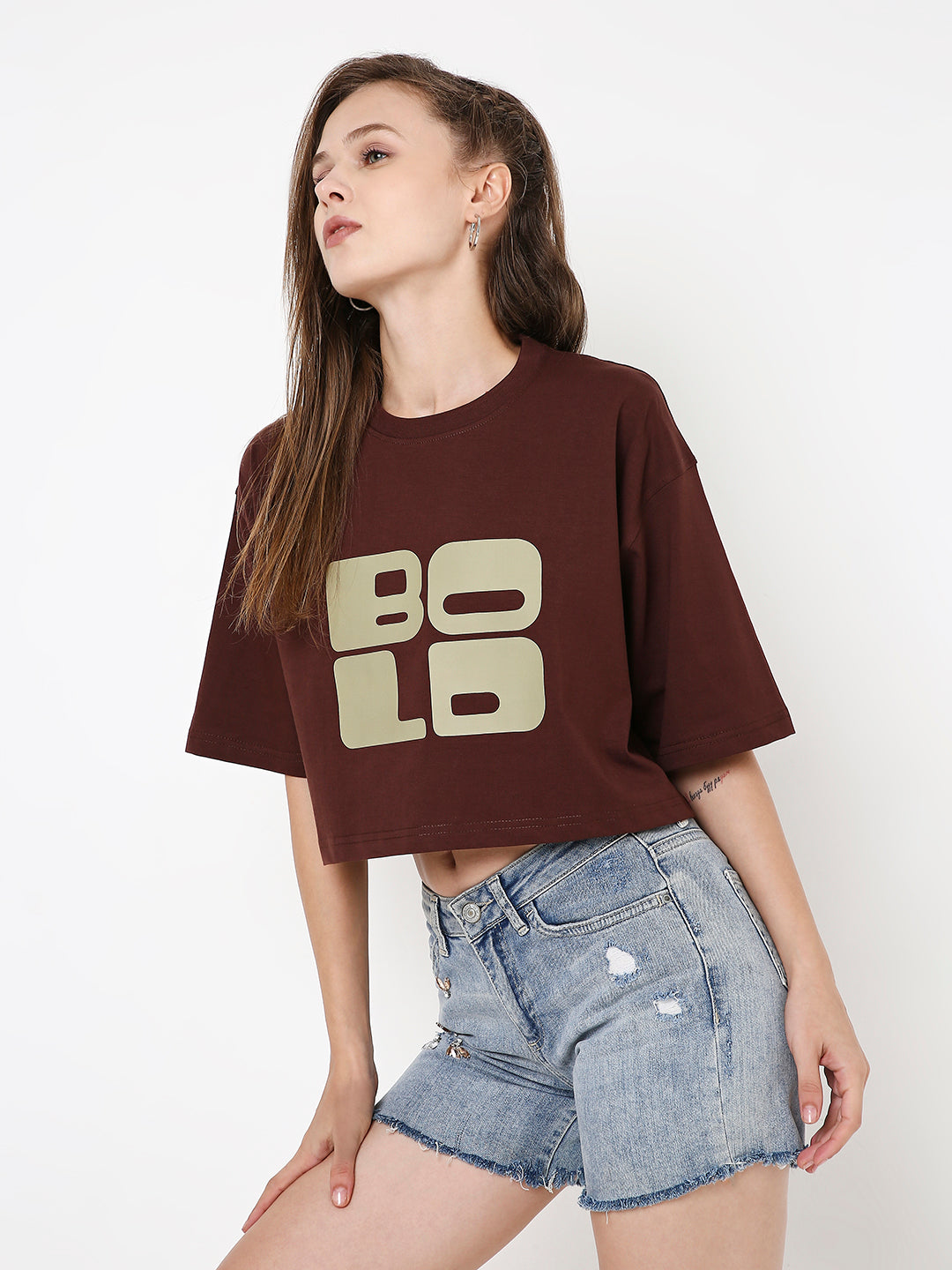 Brown oversized cropped T-shirt with bold beige "BOLD" print, paired with distressed denim shorts.