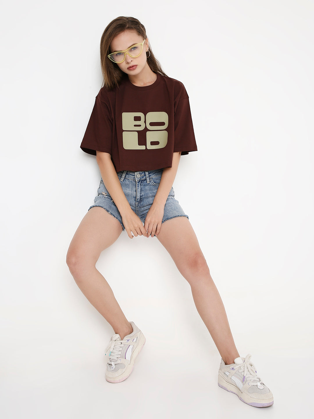 Oversize brown crop top with bold beige "BOLD" print, paired with denim shorts.