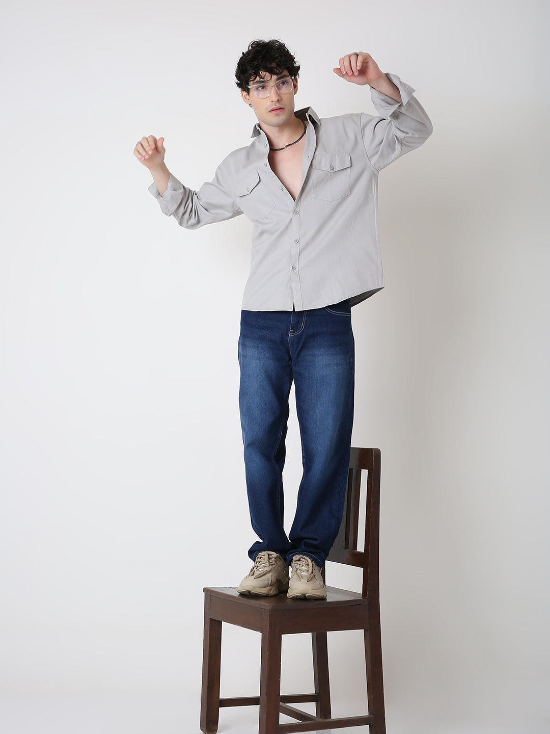 Man standing on a chair wearing a light gray shirt and blue jeans against a white background