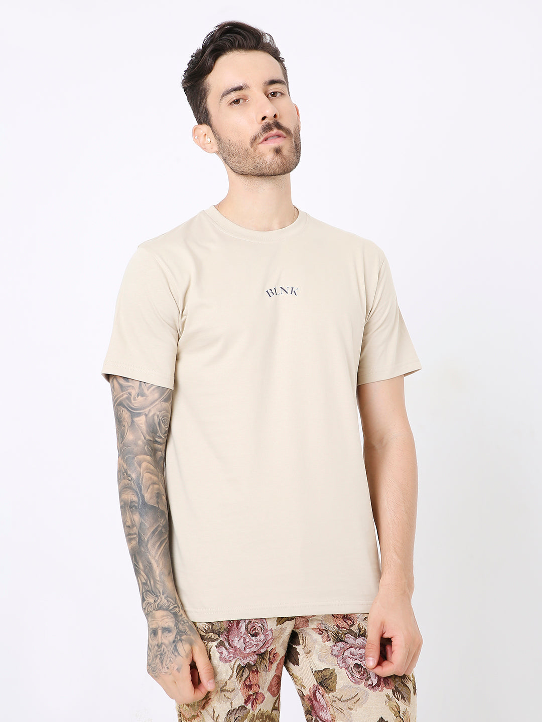 Beige relaxed fit t-shirt with small centered text on chest, worn with floral patterned pants.