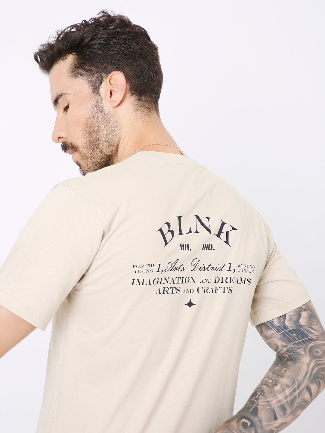 Beige relaxed fit t-shirt with black graphic text and design on the upper back, worn by tattooed male model.