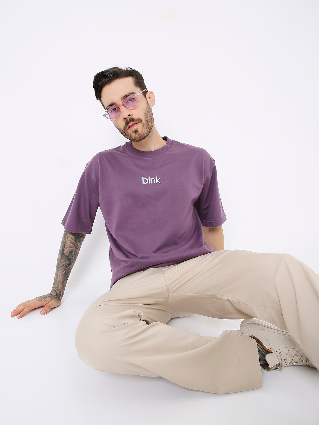 Oversize purple t-shirt with white "bink" text on chest, worn with beige wide-leg pants and tinted sunglasses.